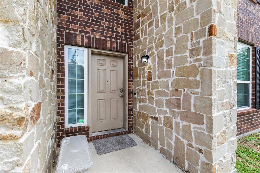 Charming entrance featuring a tan door surrounded by a mix of brick and stonework, with a small window and outdoor light fixture.