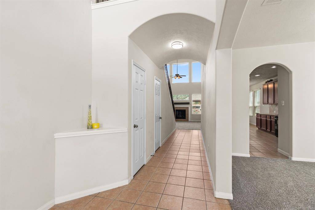 This photo showcases a spacious hallway with high ceilings, leading to a living area featuring large windows and a fireplace. To the right, there's an arched entry to a kitchen with ample cabinet space. The flooring is a mix of tile and carpet, creating a warm, inviting atmosphere.