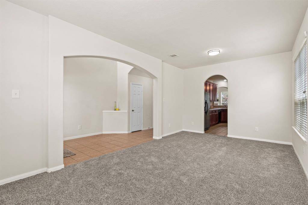 This photo showcases a bright, open living space with neutral walls and carpeting. It features arched doorways leading to a tiled area and a modern kitchen with dark cabinetry. Large windows provide ample natural light.