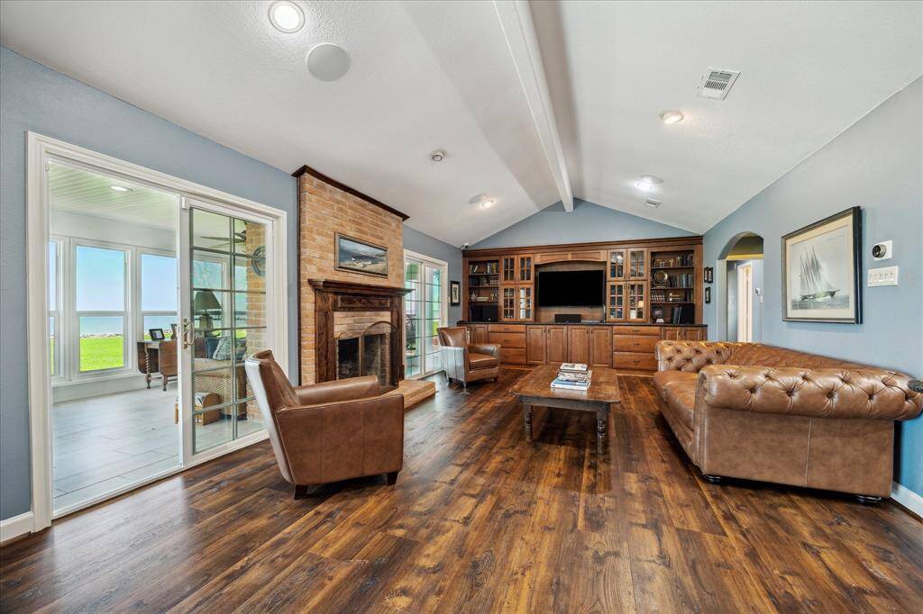 A view into the den. At the left, on each side of the fireplace, are slider doors that provide a great view of the water and access to the sunroom. The den has a beamed vaulted ceiling, recessed lighting, double slider doors to the sunroom and backyard, and is wired for sound. At the far end is a built-in entertainment center with granite surface that includes upper glass front cabinets, open shelving and lower drawer and cabinet storage.