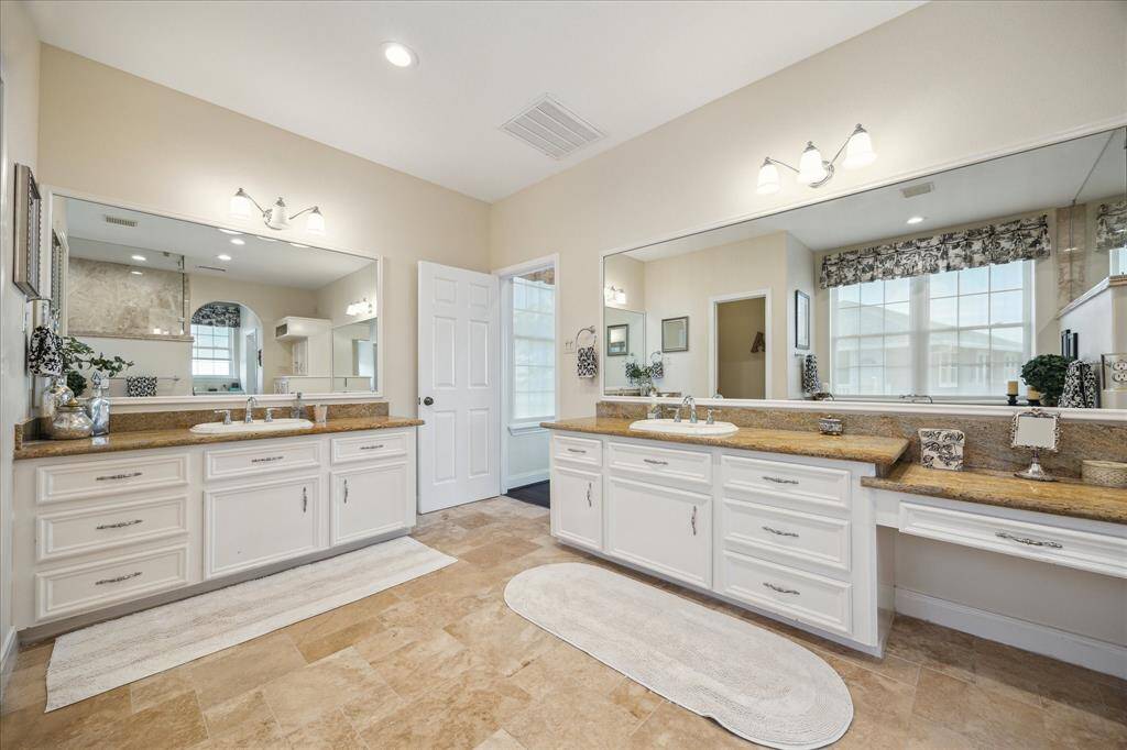 The en-suite primary bath has travertine floor, split vanities with granite tops, one with a dressing area, and custom vanity width mirrors.