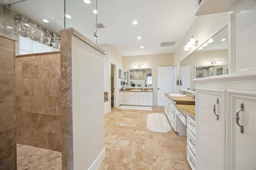 This view across the primary bath shows the entrance to the separate shower that has travertine surround with stone accent, built-in shelving, rain shower and frameless glass.