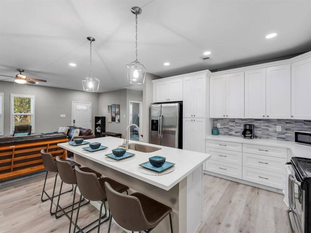 This open-concept kitchen features modern white cabinetry, a sleek island with seating, stainless steel appliances, and stylish pendant lighting. The space flows into a cozy living area with natural light from large windows. Perfect for entertaining and everyday living.
