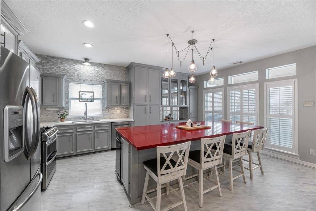 This (2018) modern kitchen features a striking red island with seating, sleek gray cabinetry, and stainless steel appliances. The space is brightened by ample natural light from large windows and stylish pendant lighting.