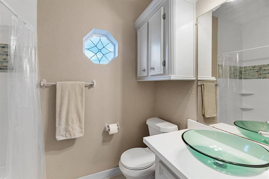 This bathroom features a modern design with a glass vessel sink, sleek white cabinetry, and a decorative octagonal window. The shower area has stylish tile accents, complementing the light and airy atmosphere.