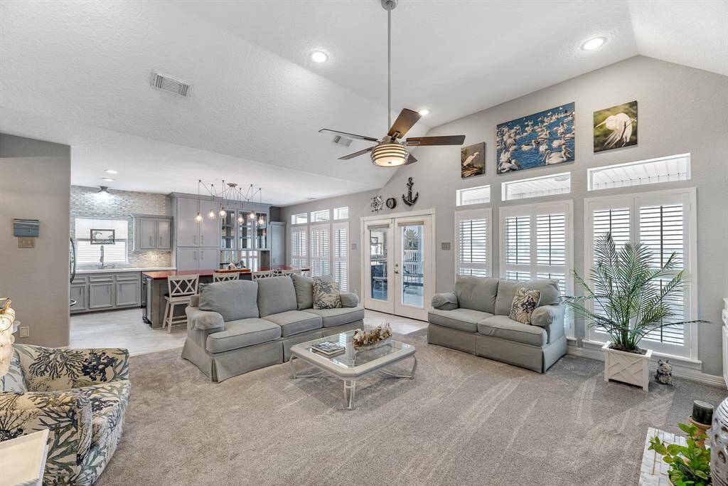 This inviting living space features an open-concept design with vaulted ceiling, abundant natural light, stylish gray cabinetry, and a cozy seating area. The room is enhanced by a modern ceiling fan, and elegant plantation shutters, creating a warm and welcoming atmosphere perfect for relaxation and entertainment.