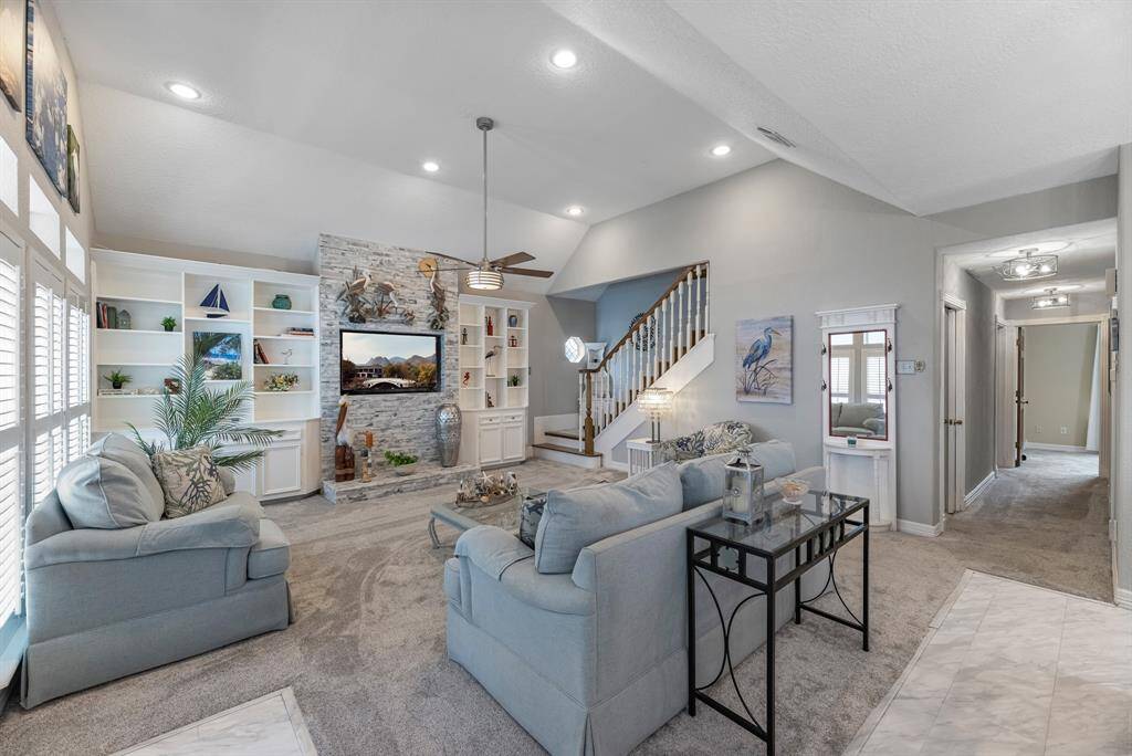 Spacious living room with high ceilings and abundant natural light. Features built-in shelving, a stone accent wall equipped with a TV space. Includes modern lighting, and a neutral color palette.