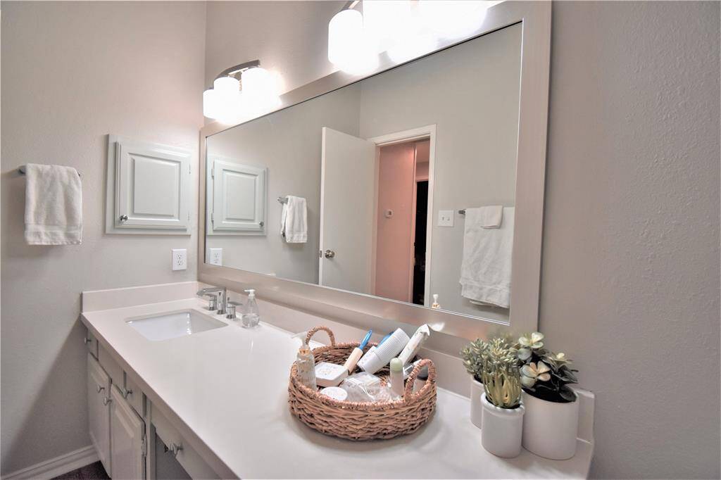 Wide mirror with wall mounted light, white countertops and cabinetry.
