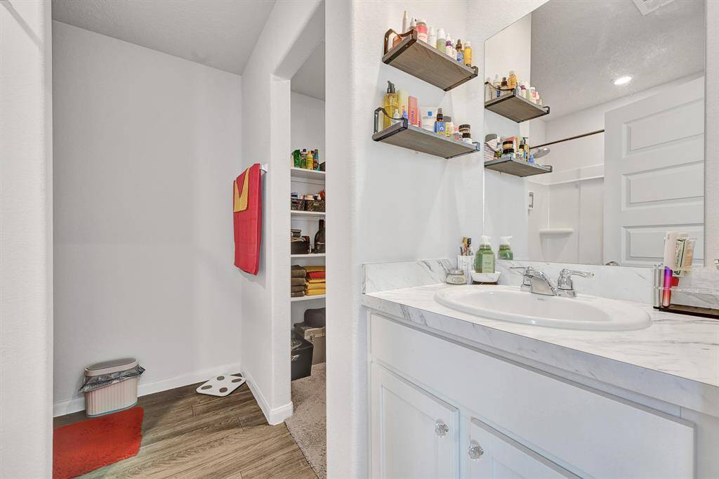 Modern primary bathroom with white cabinetry and a marble look countertop. It features a sink with dual faucets, open shelving for toiletries, and a glimpse of a walk-in closet with organized shelving for storage. The space is bright and functional, ideal for everyday use.