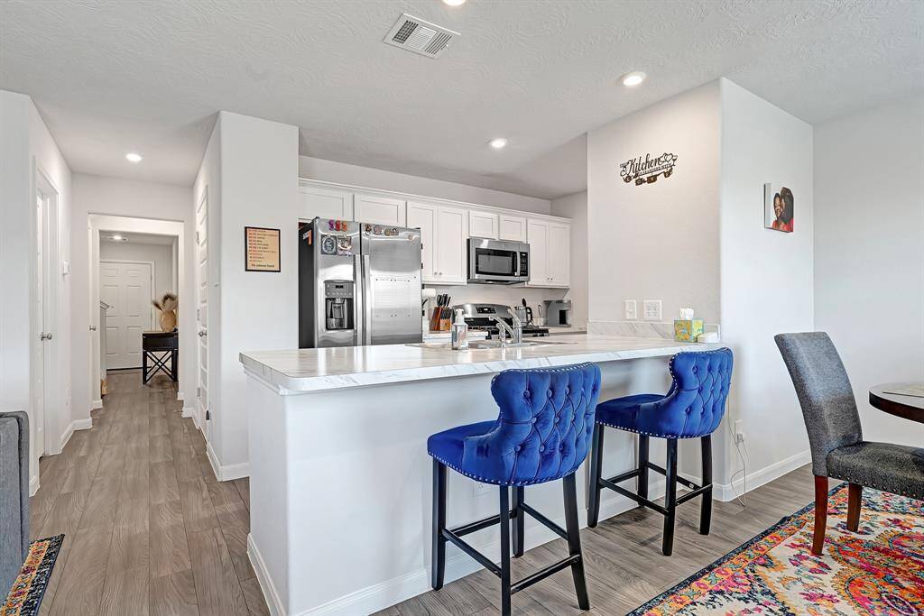 The modern kitchen offers white cabinetry, stainless steel appliances, and a sleek countertop. The space includes a breakfast bar with seating space, easy to clean flooring, and ample lighting, creating a bright and inviting atmosphere.
