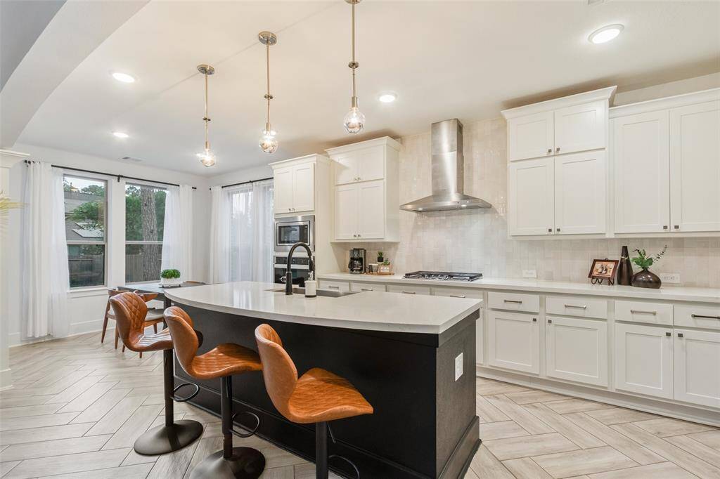 This modern kitchen features sleek white cabinetry, a large island with seating, and stainless steel appliances. It has ample natural light from large windows, pendant lighting, and a stylish range hood, creating a bright and inviting space.