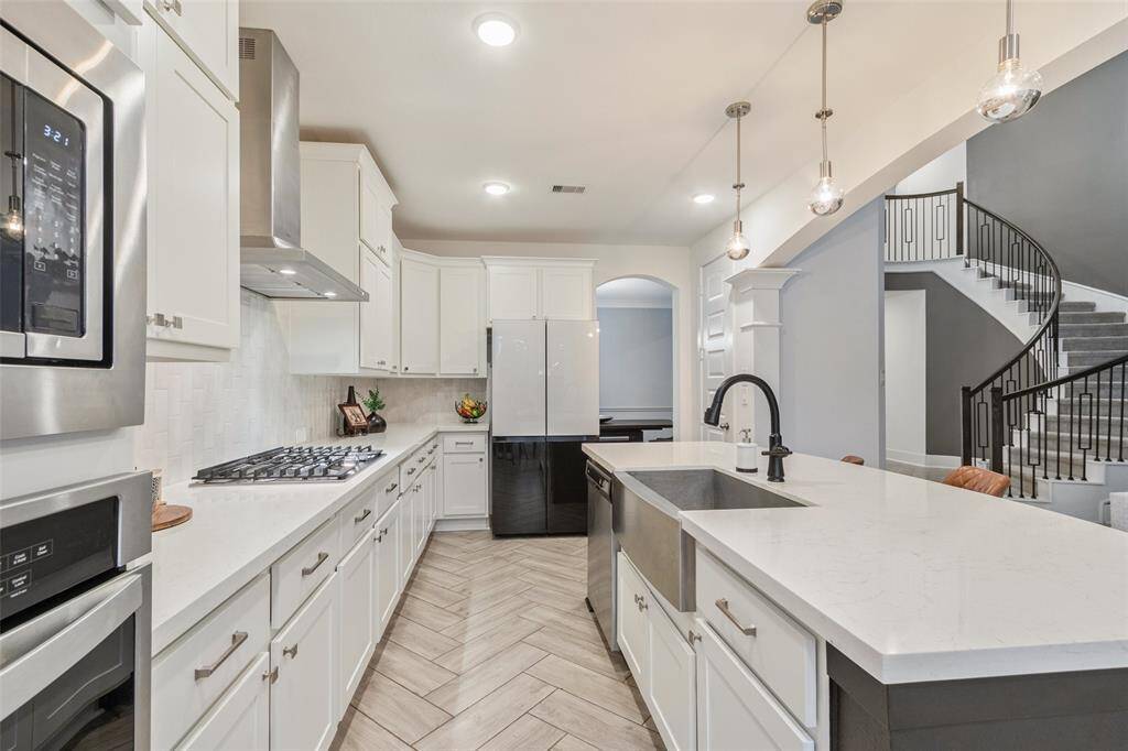 This modern kitchen features a spacious island with a farmhouse sink. Elegant pendant lighting and herringbone-patterned flooring enhance the contemporary design, while an open layout seamlessly connects to a grand staircase.