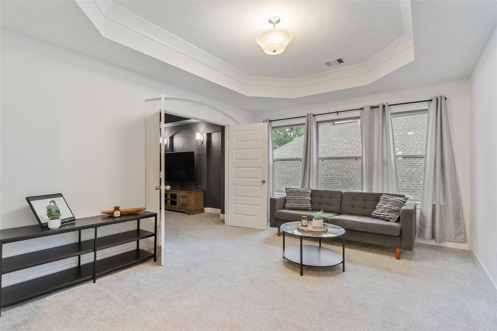 This bright, spacious room features a cozy sitting area with  large windows, offering plenty of natural light. The neutral carpet and tray ceiling add elegance, while an open doorway leads to a media room, perfect for entertainment.