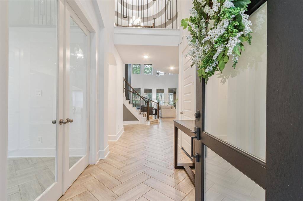 This inviting entryway features elegant herringbone tile flooring, a grand staircase with wrought iron railing, and abundant natural light. The double glass doors on the left lead to a spacious room, while a decorative wreath adorns the front door, adding a welcoming touch.