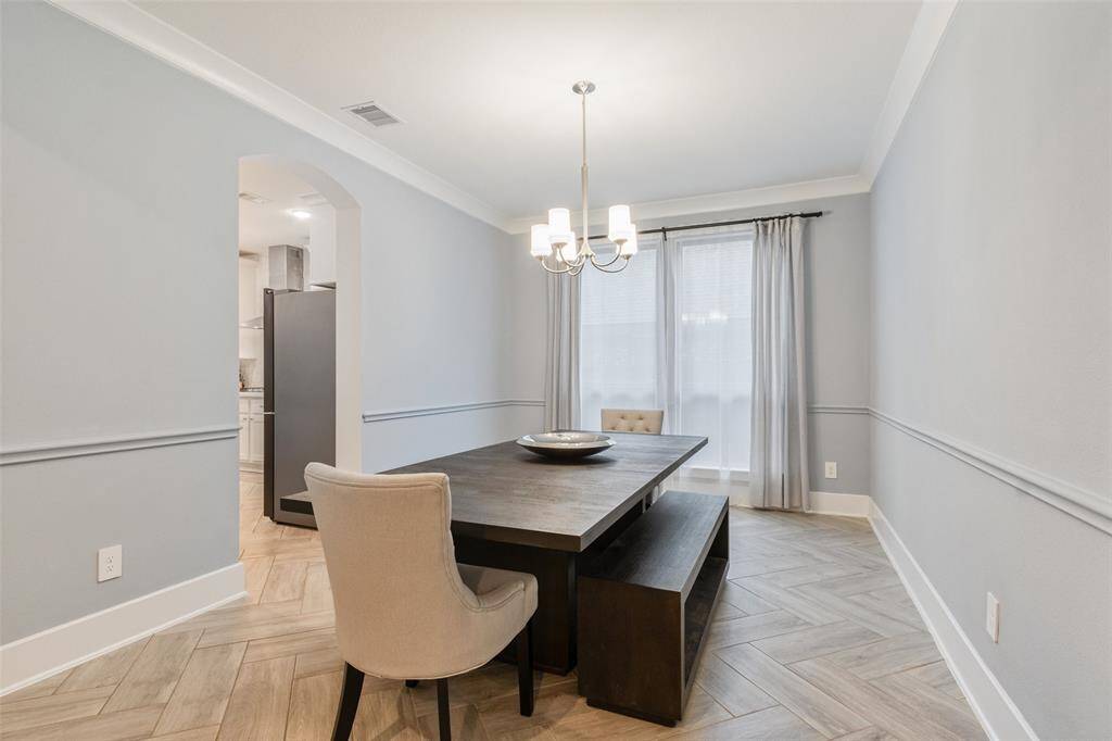 This photo shows a modern dining room with light gray walls and herringbone-patterned flooring, and  features  a stylish chandelier. A large window with sheer curtains provides natural light, and an arched opening leads to the kitchen.