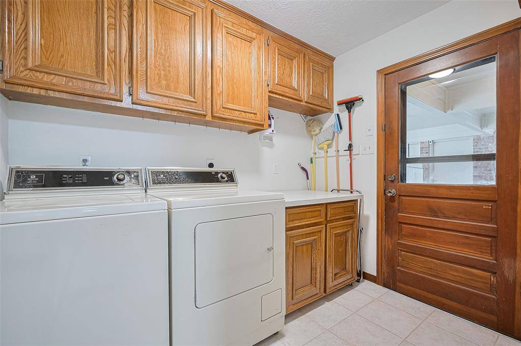 The utility room, measuring 8'8" x 5'10", includes a full-size Maytag washer and dryer, with both electric and gas dryer connections for flexibility. Upper storage cabinets offer organization, and a quartz countertop provides a convenient space for folding clothes. The cabinet could be removed to accommodate a second refrigerator or upright freezer, if desired. Door on the right leads to the covered patio, porte corchere, garage, and backyard.