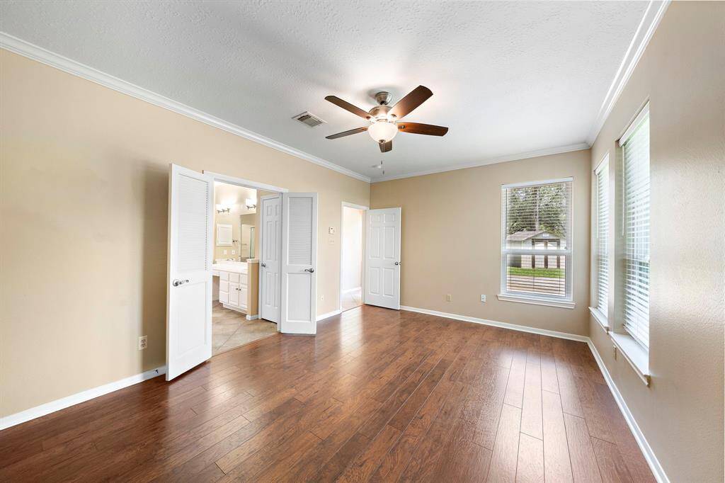 This bright, spacious primary room features hardwood floors, neutral walls, and a ceiling fan. Large windows provide natural light, and doors lead to the adjoining en-suite bathroom with dual sinks.
