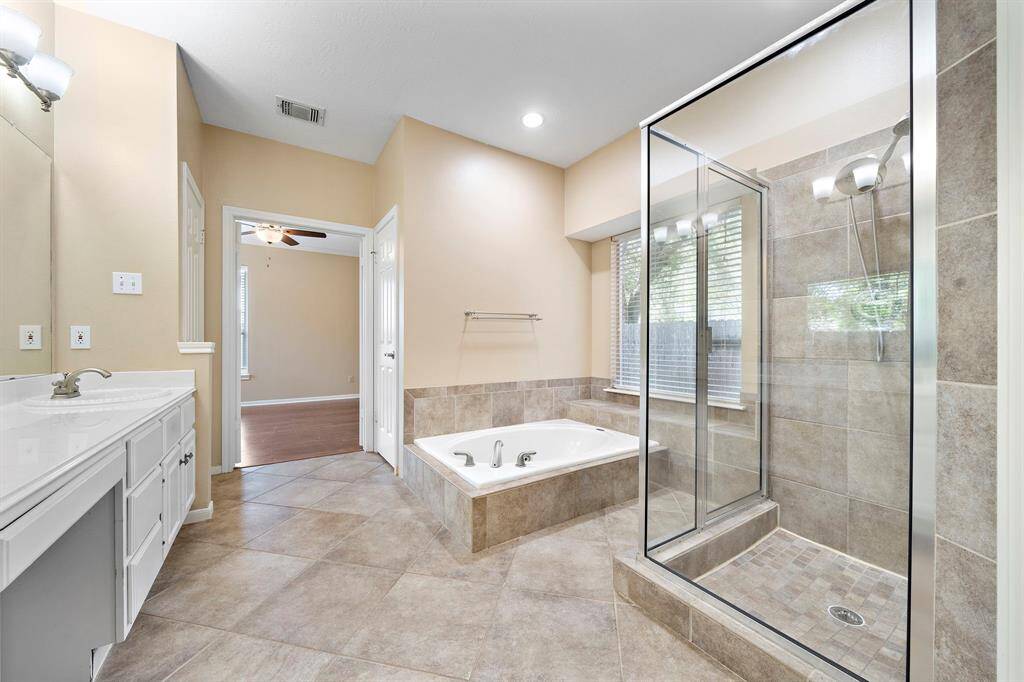 This spacious bathroom features a large glass-enclosed shower, a separate bathtub, dual sinks with ample counter space, and neutral tile flooring. Natural light filters through a window, enhancing the room's warm ambiance.