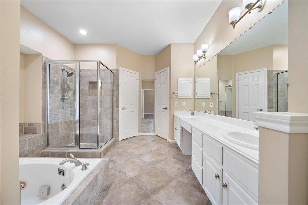 Spacious primary bathroom featuring a large glass-enclosed shower, a soaking jetted tub, and dual sinks with ample counter space. Neutral-toned tiles and a wide mirror create an elegant and inviting atmosphere.