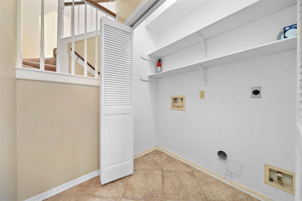 This photo shows a laundry area with hookups for a washer and dryer, open shelving for storage, and a tiled floor located just outside the primary bedroom across from the powder room.