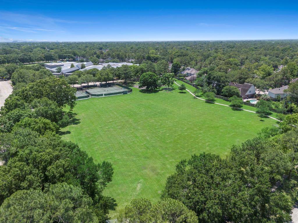 Expansive green park surrounded by trees and residential homes, featuring a tennis court area ideal for outdoor activities and community living.