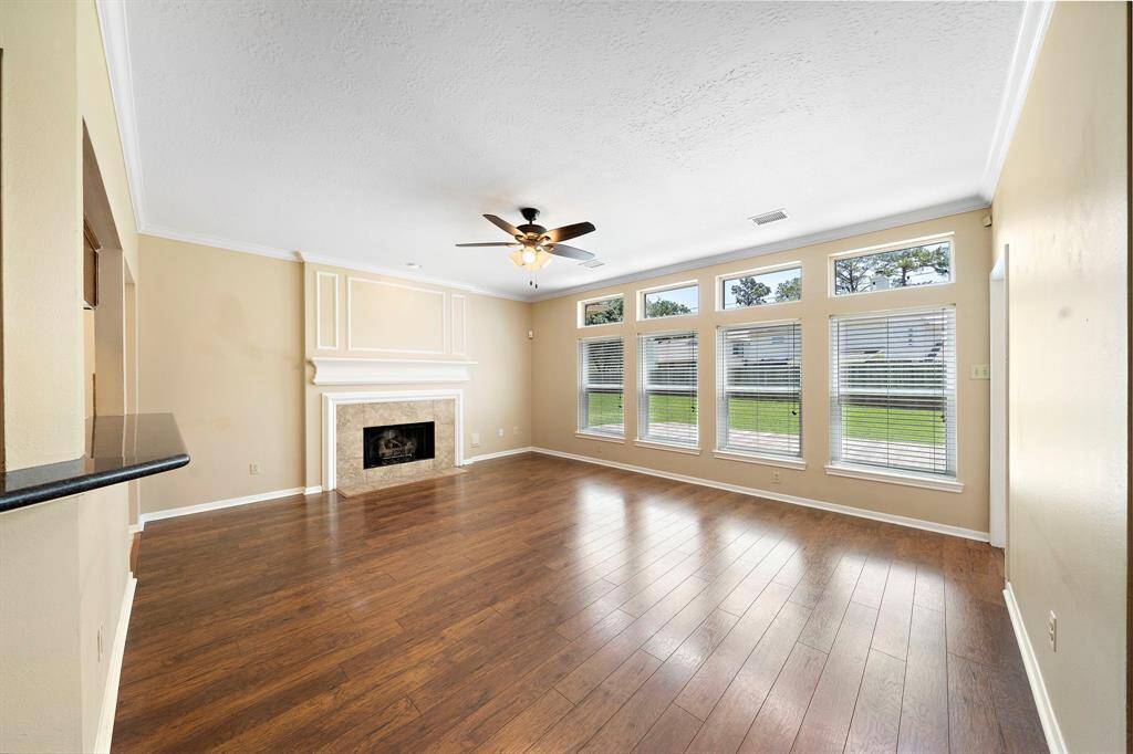 The main living room features a cozy fireplace, elegant wood flooring, and abundant natural light from large windows overlooking the backyard. A ceiling fan adds comfort, and the neutral walls offer a welcoming and versatile backdrop.