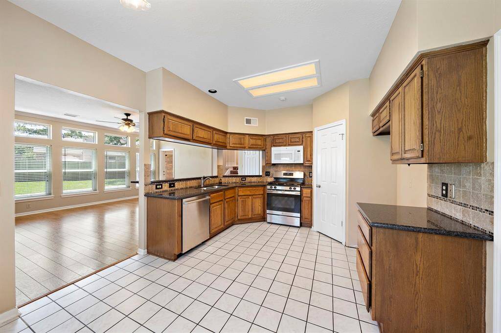 This photo showcases a bright, open kitchen with wooden cabinets, granite countertops, and modern stainless-steel appliances. It features tile flooring and opens up to a spacious living area.