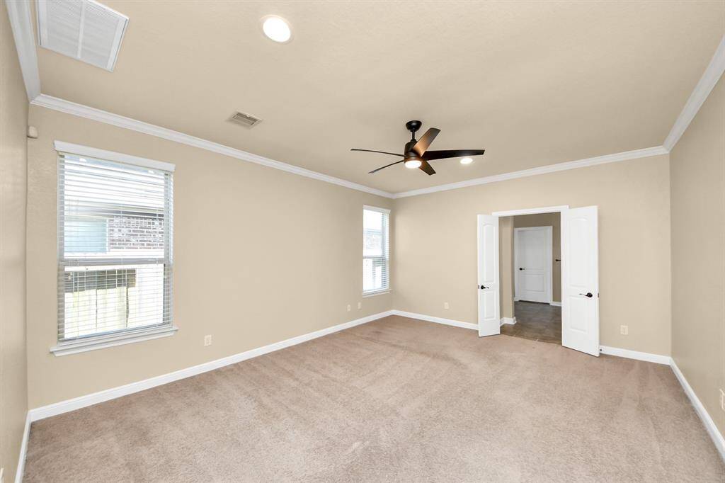 This room features soft beige walls and carpet flooring, offering a warm and neutral space. It includes two large windows for natural light, a modern ceiling fan, and double doors leading to an adjacent area.