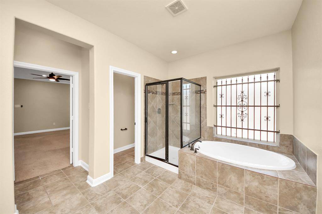 This spacious bathroom features a tiled floor, a glass-enclosed shower, and a large soaking tub with decorative window bars. Neutral tones create a calm atmosphere, and it connects to a carpeted room.