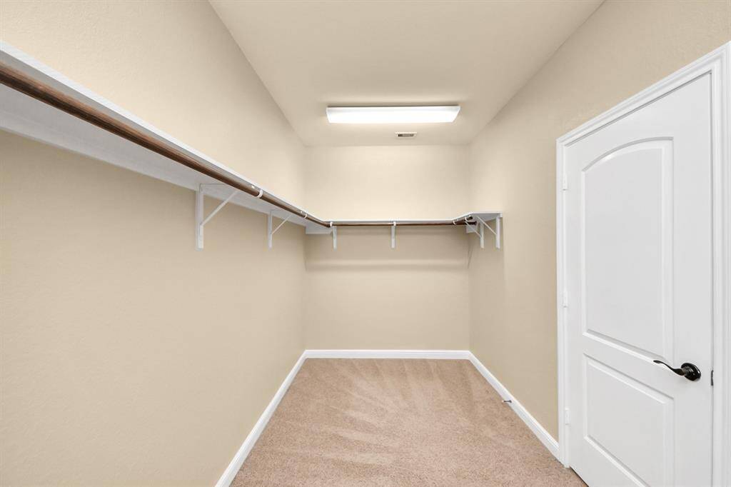 This photo shows a spacious walk-in closet with beige walls, carpeted flooring, and ample shelving for storage. A white door and ceiling light complete the clean, bright look.