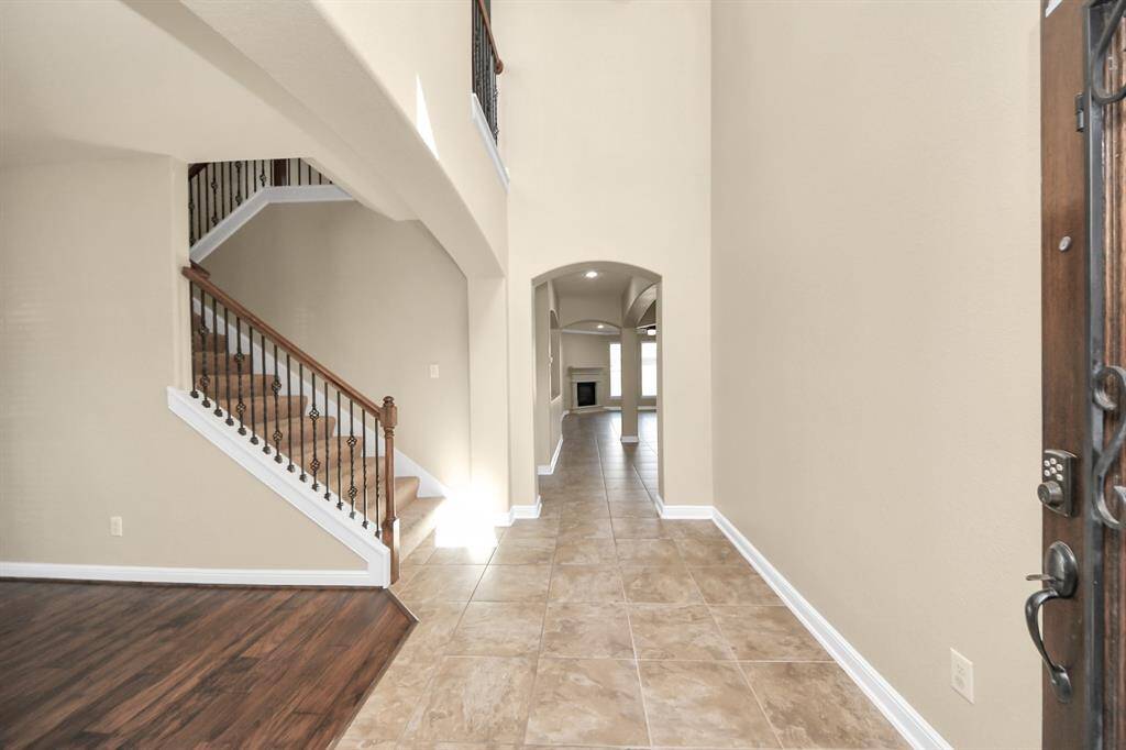 This photo showcases a spacious entryway with tiled flooring and high ceilings. There's a staircase with wooden handrails and wrought iron balusters to the left. The hallway leads to a well-lit living area with a fireplace, visible through an arched opening.