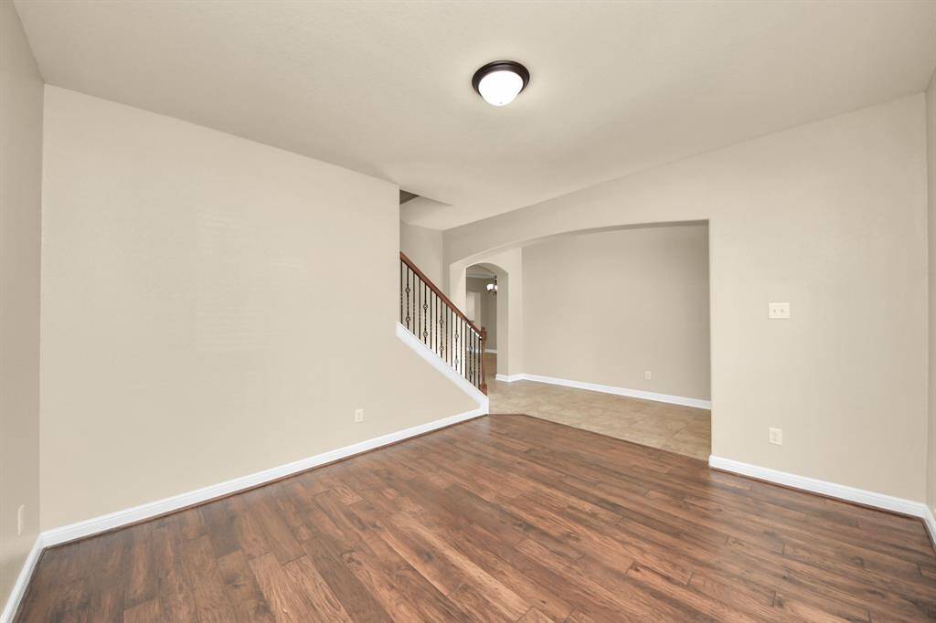This photo showcases a spacious room with wood flooring and neutral walls, leading to a stairway with a wooden handrail. It features an arched entryway to a tiled area, offering a modern and open feel.