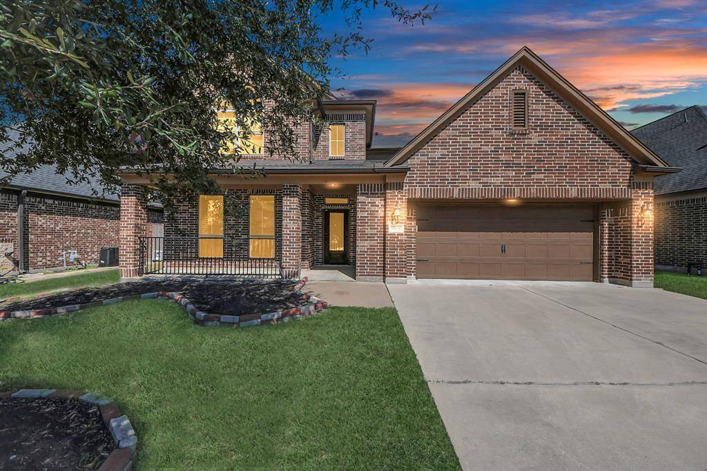 This inviting brick home features a two-car garage, well-maintained front yard, and charming porch. It's enhanced by warm, welcoming lighting and set against a beautiful sunset backdrop.