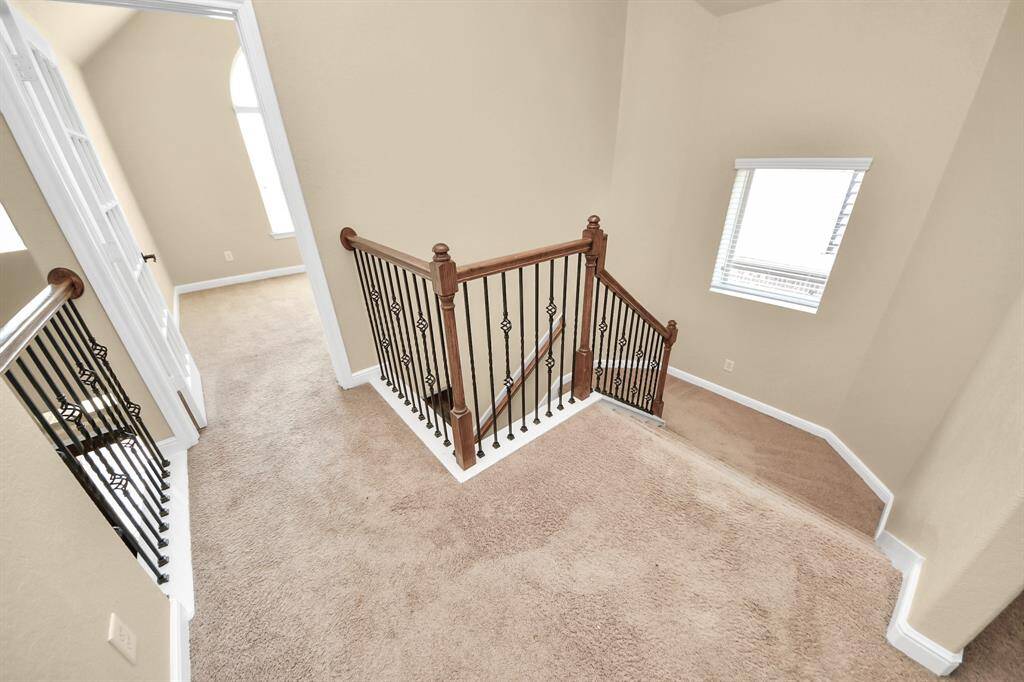 This photo showcases an upstairs landing area with beige carpeting and elegant wrought iron railings. The space is brightened by a window, leading to rooms on either side, and features neutral wall colors.