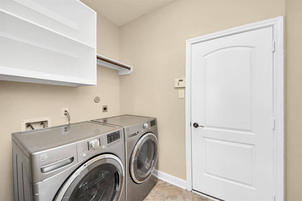 This photo shows a modern laundry room featuring a front-loading washer and dryer. There's a white door, beige walls, and overhead shelving for storage. The space is clean and functional.