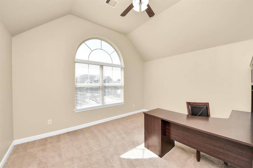 This is a bright, spacious room with a vaulted ceiling and large arched window, providing ample natural light. It features a neutral color scheme, carpeted floor, and includes a modern desk, ideal for a home office or study.