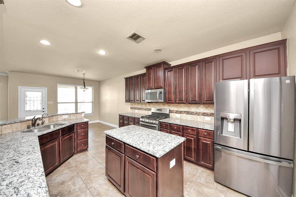 Spacious kitchen with dark wood cabinetry, granite countertops, and stainless steel appliances. Features a center island, ample lighting, and a breakfast nook with large windows. Ideal for cooking and entertaining.
