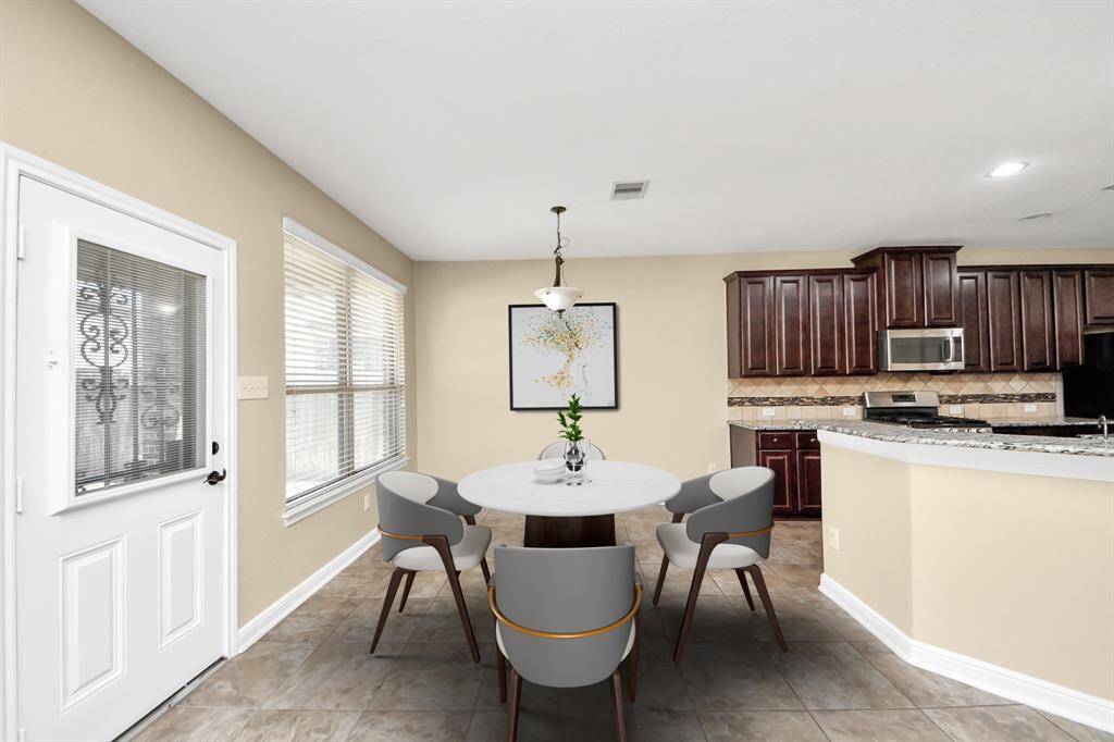 Bright dining area with tile flooring and a round table, adjacent to a modern kitchen featuring dark wood cabinets and stainless steel appliances. Large windows and a door provide ample natural light. VIRTUAL IMAGE