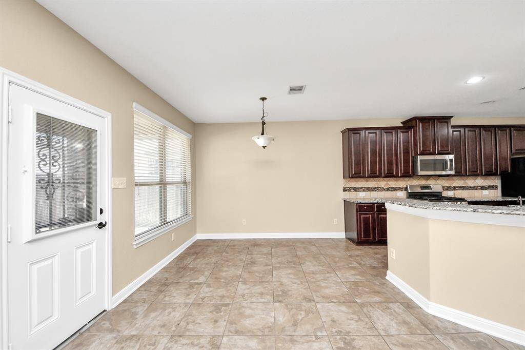 This photo shows a bright, open kitchen and dining area with beige walls and tile flooring. It features dark wood cabinetry, stainless steel appliances, and a granite countertop. There is ample natural light from large windows and a door with glass inserts.