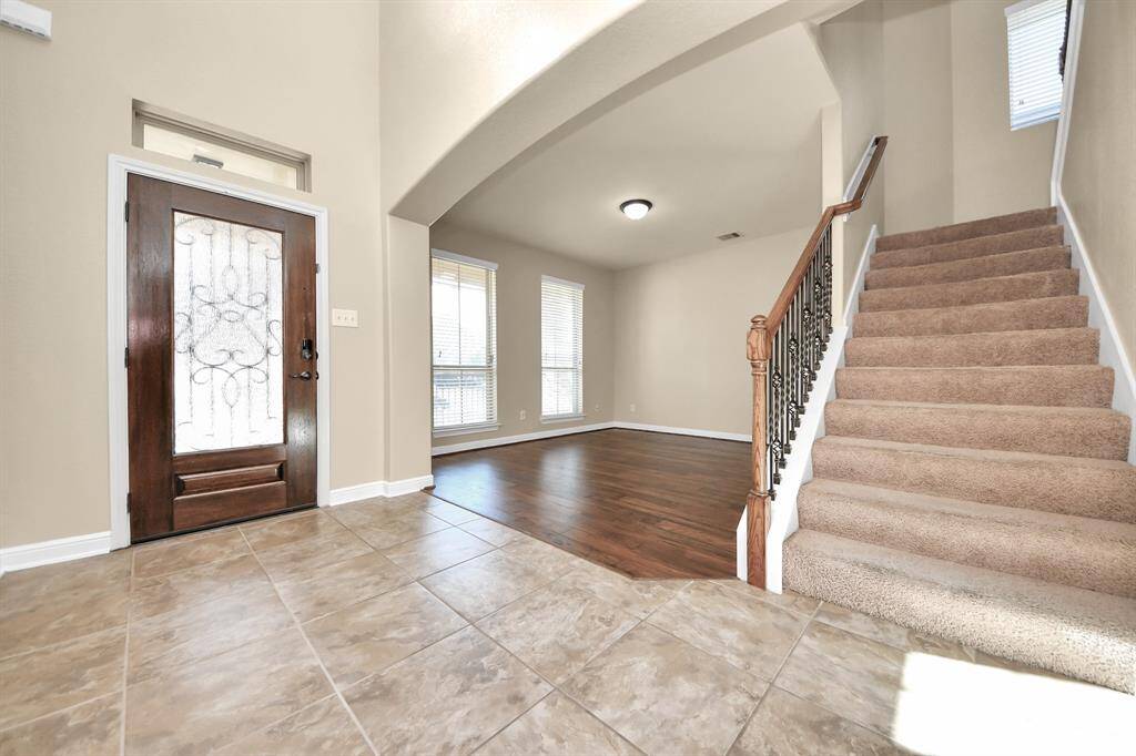 This welcoming entryway features a decorative front door, tile flooring, and a carpeted staircase. The room opens to a bright living area with hardwood floors and large windows, creating a warm and inviting atmosphere.