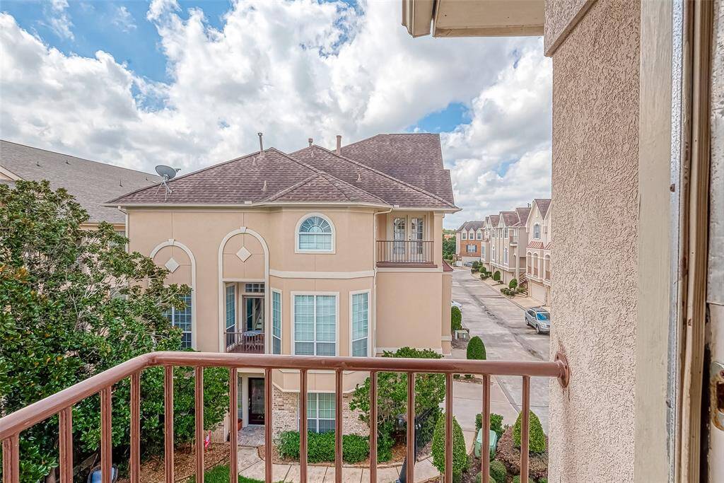 This photo showcases a view from a balcony overlooking a charming residential street. The neighborhood features well-maintained homes with a classic design, surrounded by greenery. The sky is bright and partly cloudy, enhancing the inviting atmosphere.