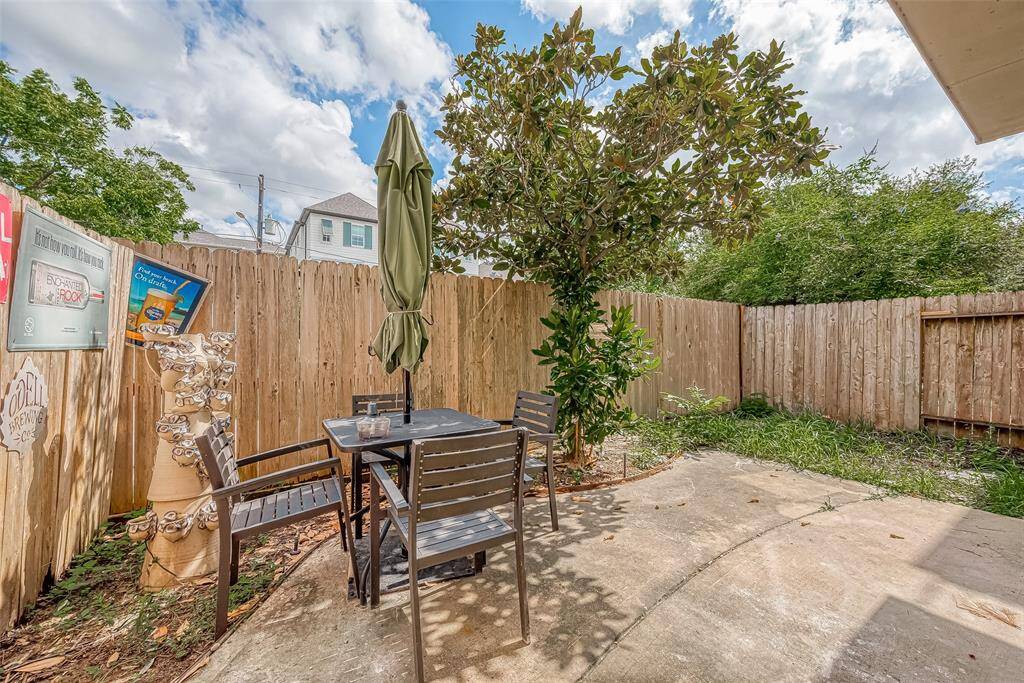 This cozy backyard features a small patio area with a table, chairs. surrounded by a wooden fence for privacy. A tree provides natural shade, and paved surfaces, perfect for outdoor relaxation.