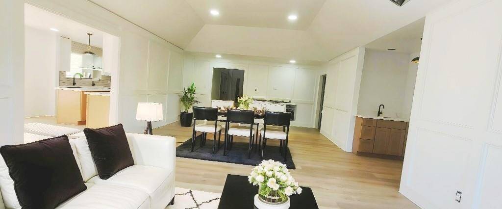 This photo showcases a spacious, open-concept living and dining area. It features light wood flooring, white walls, and recessed lighting. A cozy sitting area with a white sofa and a sleek dining table are visible, along with a glimpse into a contemporary kitchen. The space is bright, welcoming, and ideal for entertaining. Virtually staged