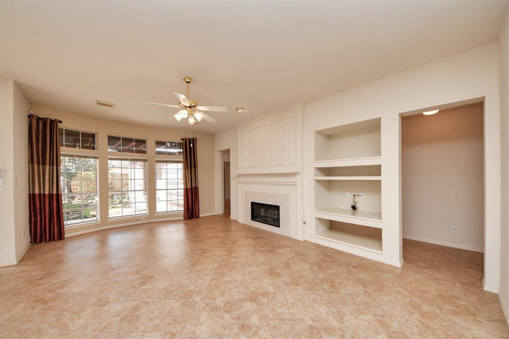The room is spacious with large windows providing plenty of natural light. It features tile flooring, built-in shelves, and a fireplace. The ceiling fan and neutral walls create a cozy atmosphere, and the space opens to a view of the backyard.