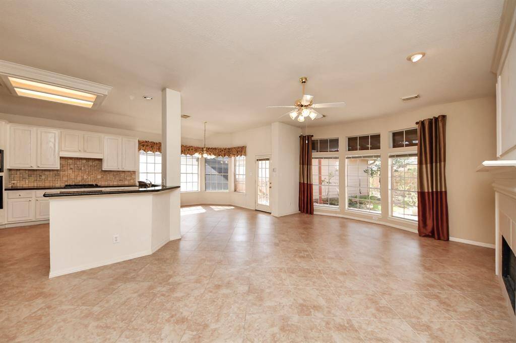The photo showcases a bright, open-concept living area with large windows, a ceiling fan, and tiled floors. The kitchen features white cabinetry, a breakfast bar, and a tiled backsplash, seamlessly flowing into the dining and living spaces.