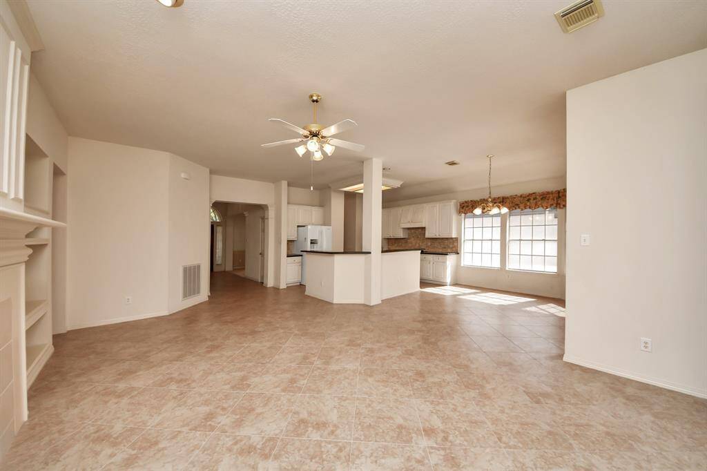This spacious, open-concept living area features tile flooring and ample natural light. It flows seamlessly into a modern kitchen with white cabinetry and a cozy dining space beneath large windows. Perfect for entertaining and family gatherings.