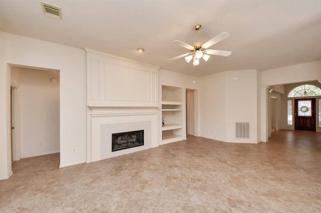 This spacious living room features a tiled floor, a central fireplace with built-in shelving, a ceiling fan, and neutral walls. The room opens to a bright entryway with a wooden front door and arched window.