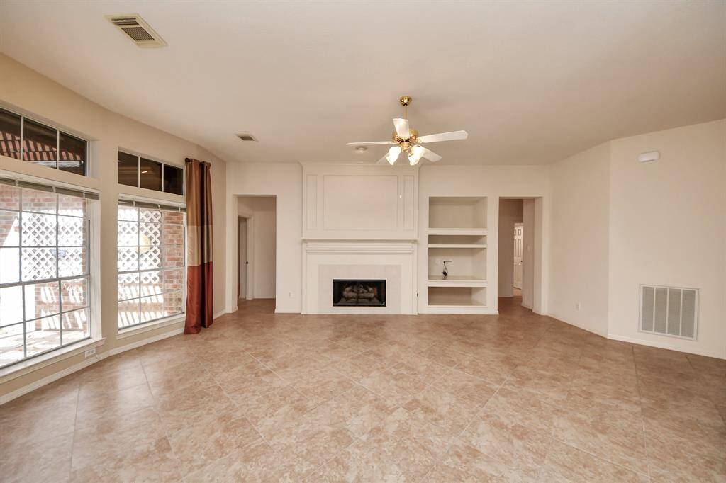 This spacious living room features large windows for natural light, tile flooring, a ceiling fan, and a cozy fireplace with built-in shelving.