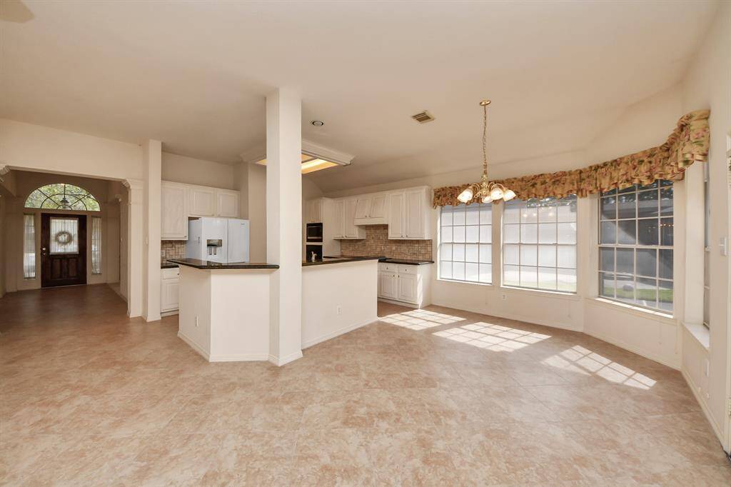 This spacious kitchen and dining area features large windows for ample natural light, white cabinetry, a modern refrigerator, and tile flooring. The open layout connects seamlessly to the entryway, creating a welcoming and airy atmosphere.