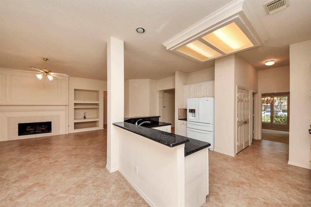 This photo shows an open-concept kitchen and living area with tile flooring. The kitchen features granite countertops and white cabinetry. The living space includes a fireplace, built-in shelving, and ceiling fan, offering a bright and spacious environment.